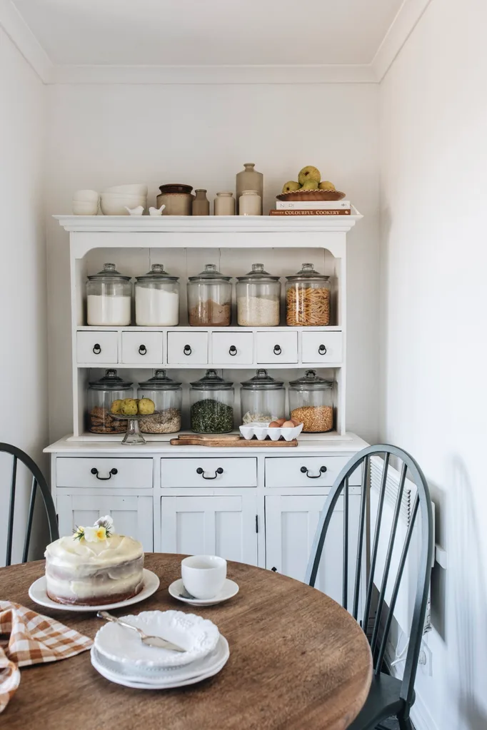 A wooden antique dining table with black chairs and a white country-style display case full of jars of pantry staples.