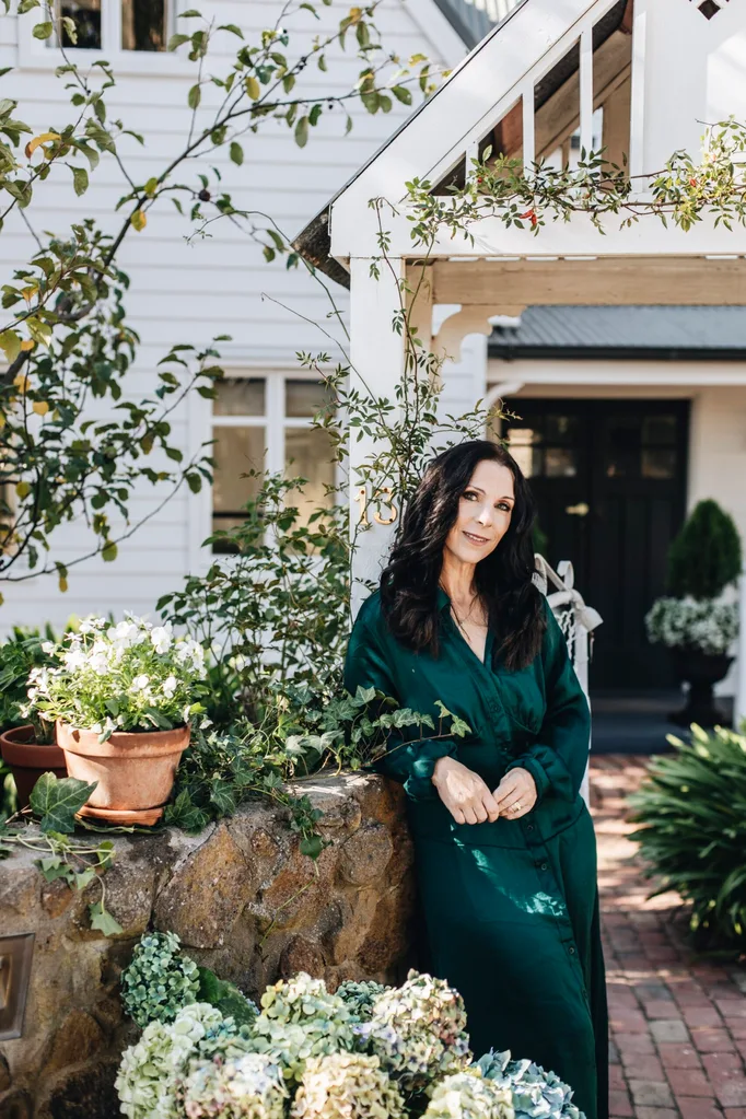 A woman in a green silk dress stands at the front of a white, storybook home.