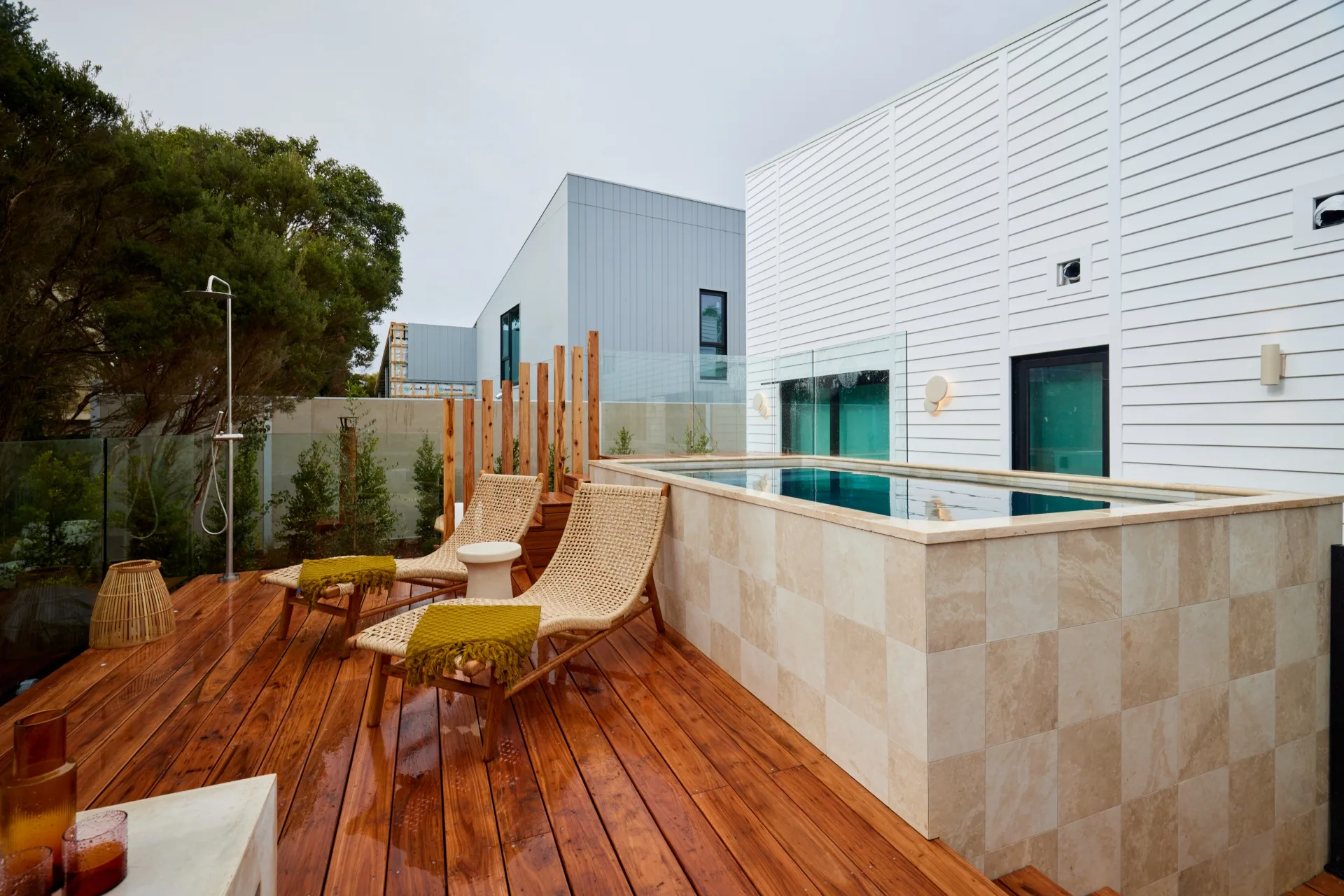 An above-ground pool with a checkerboard tile pattern, two rattan daybeds and an outdoor shower.