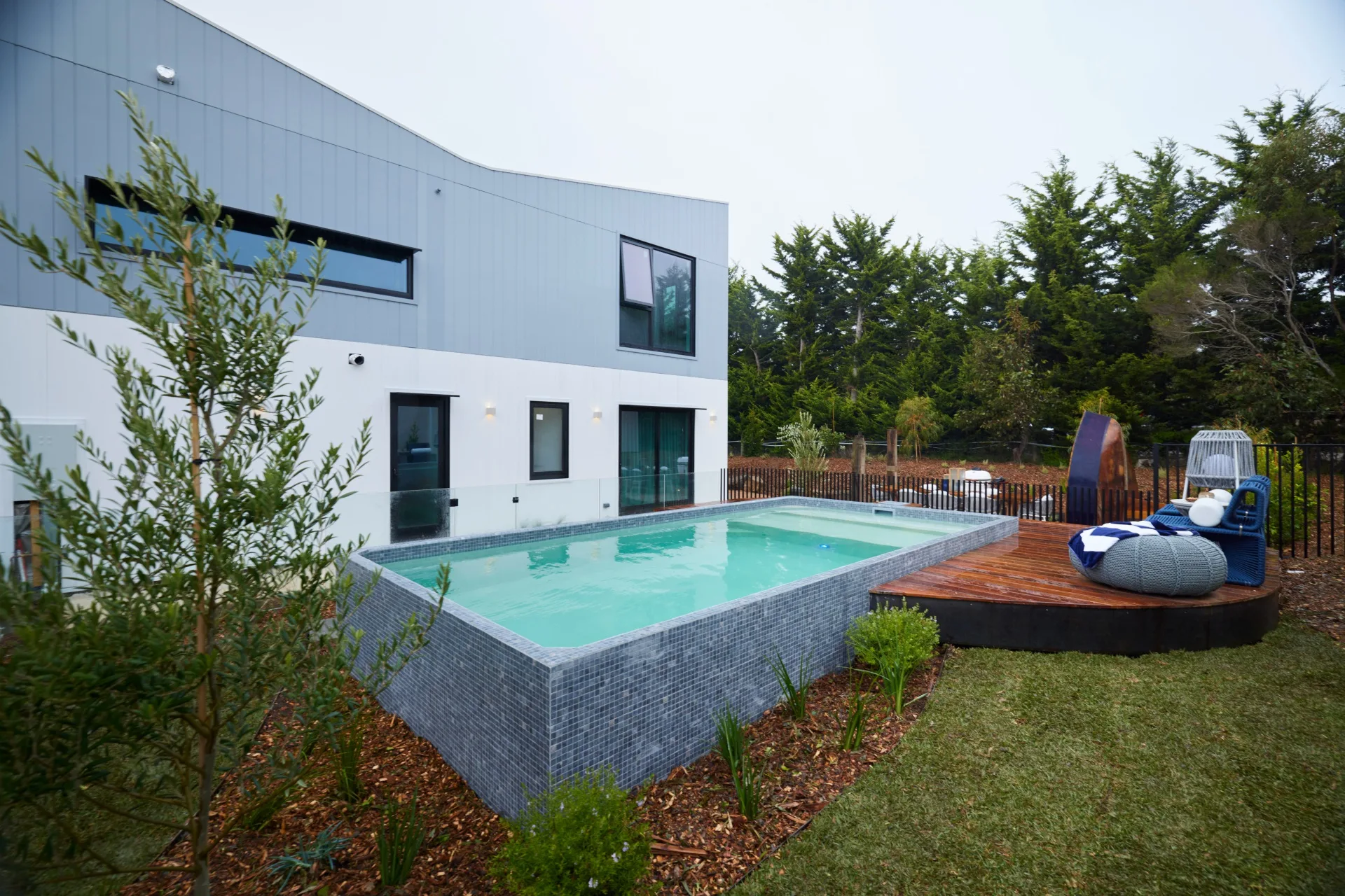 An above-ground pool with small blue tiles, a curved deck and a lawn.