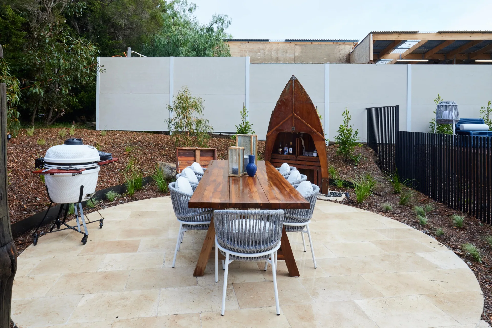 An outdoor dining area with a circular paved floor surrounded by landscaped gardens, a boat-shaped bar and a smoker.