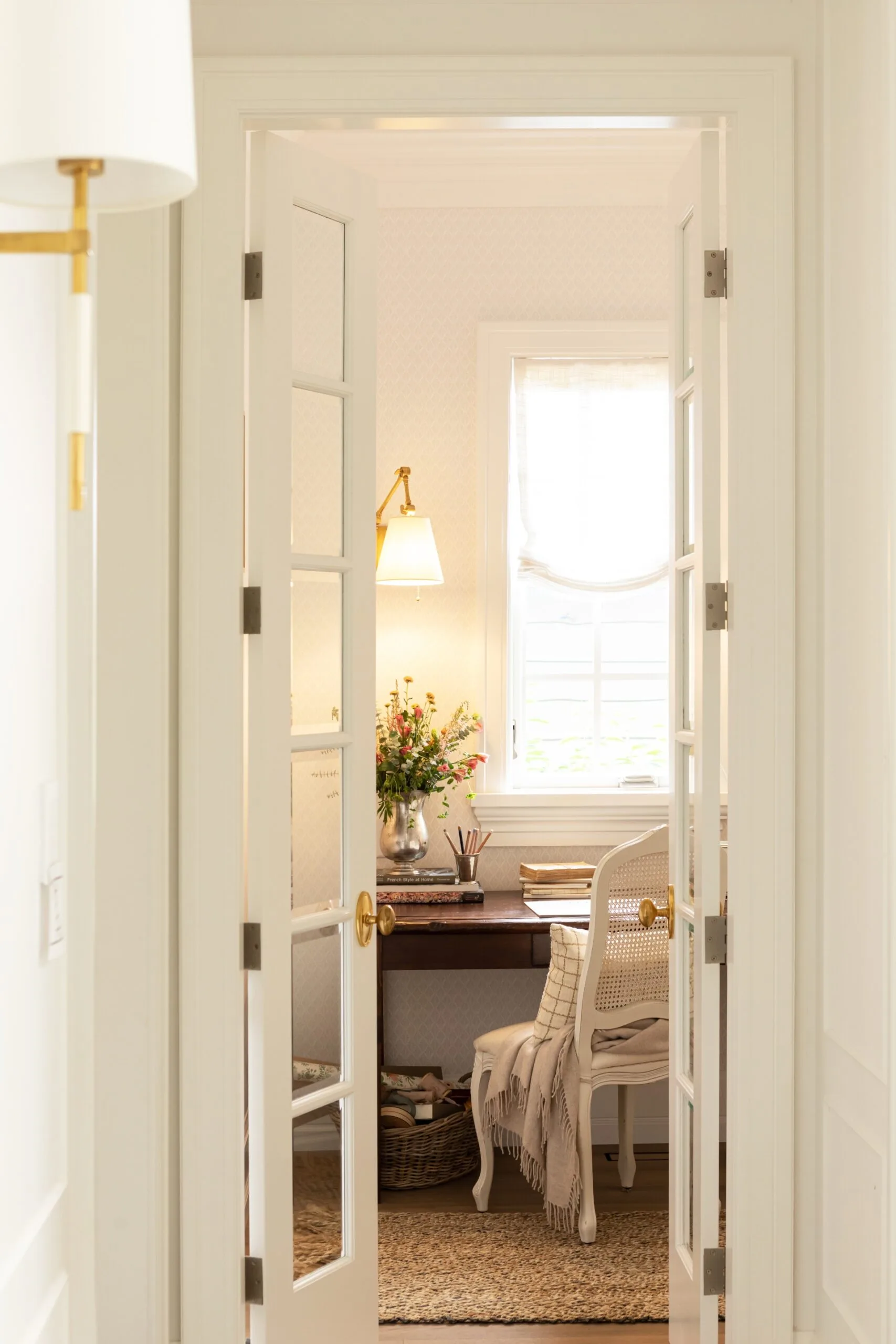 A view through two French doors to an office nook with a dark wooden table, a white antique chair and a wall-mounted sconce.
