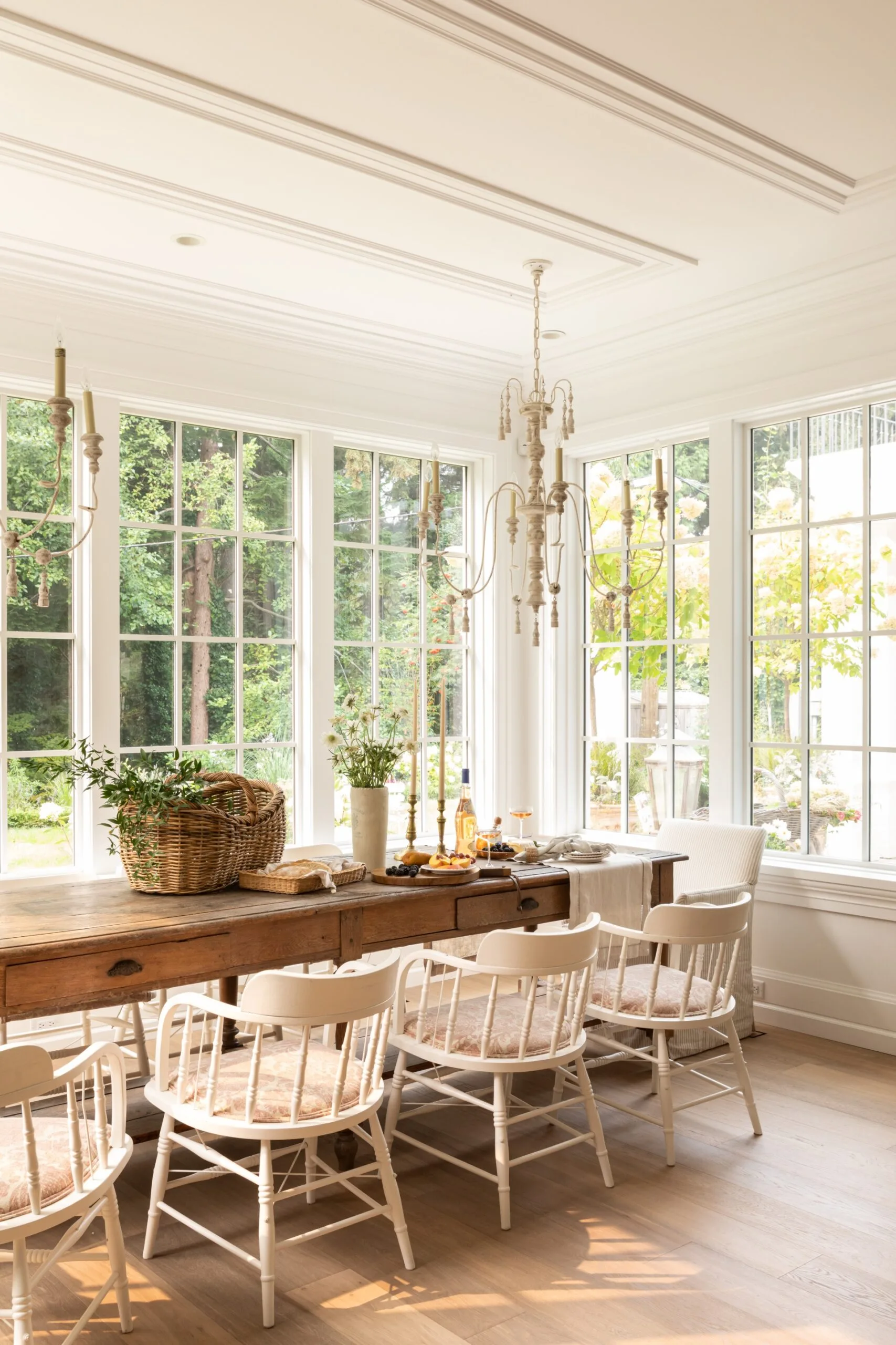 A dining room with large white windows, an antique wooden table and white chairs. A white chandelier hangs from the ceiling.