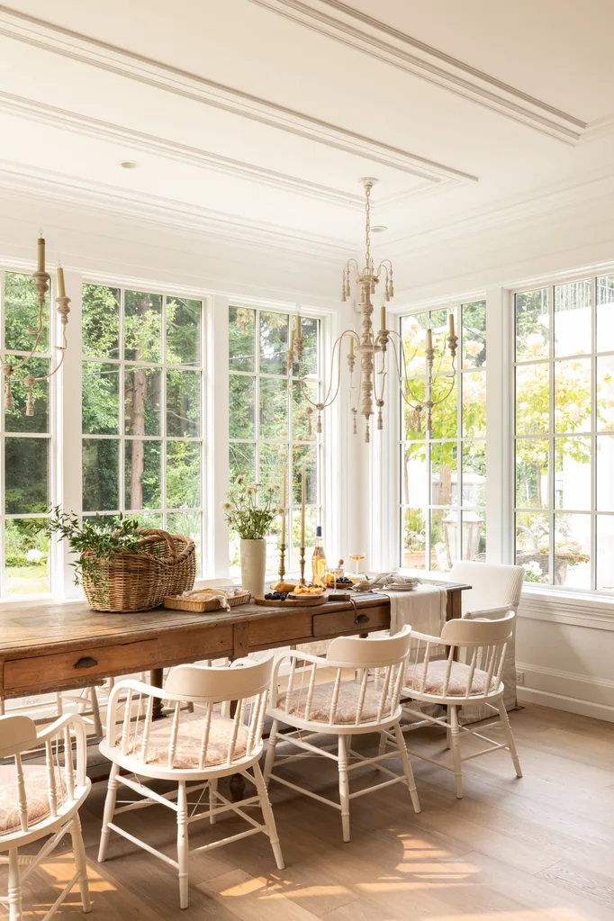 A dining room with large white windows, an antique wooden table and white chairs. A white chandelier hangs from the ceiling.