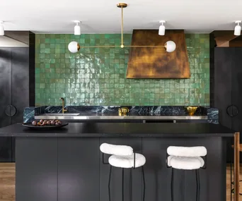 A dark kitchen with black cabinetry and a feature green splashback with a brassy rangehood