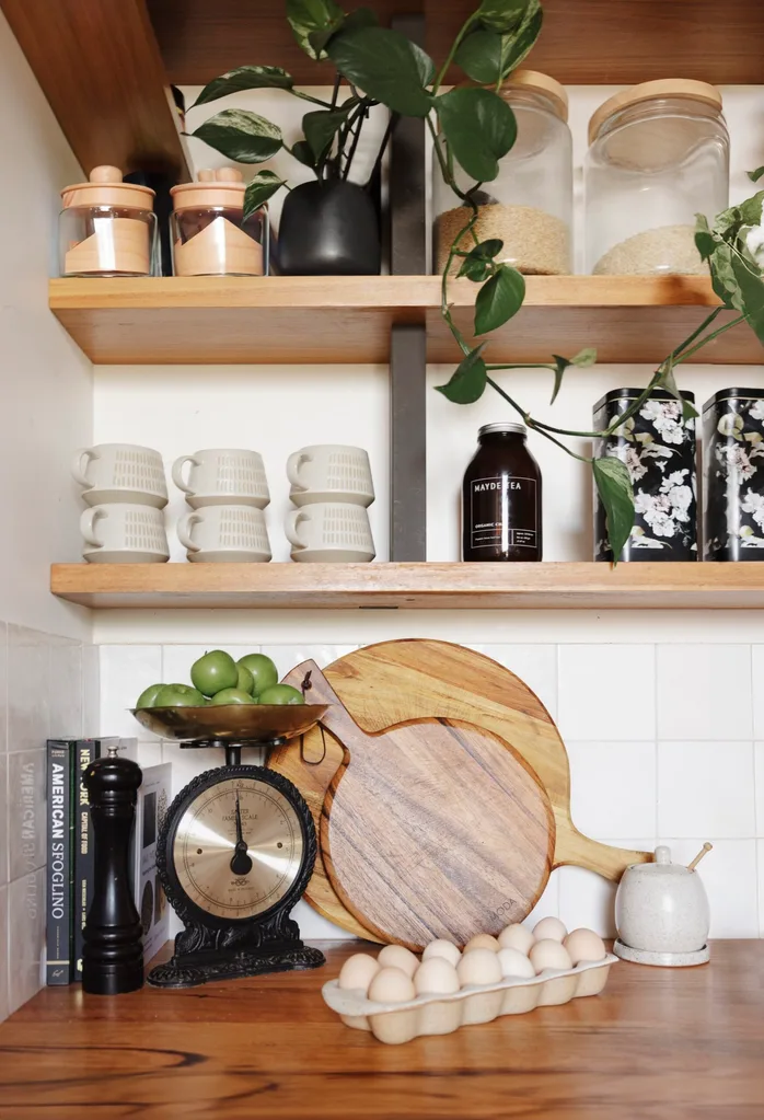 A close up of a kitchen counter topped with eggs and chopping boards; floating shelves decorated with jars and a plant hang above