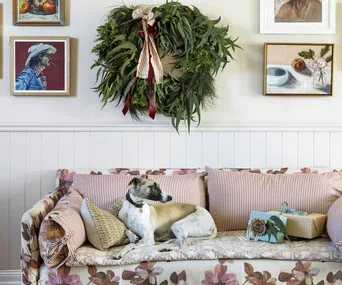 A gallery wall decorated with art and a Christmas wreath sits above a floral couch, upon which a small dog snuggles up.