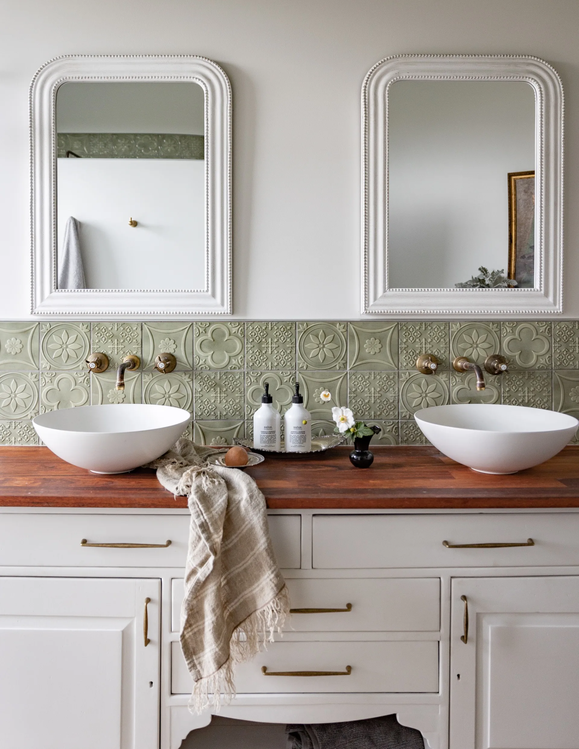 A country bathroom with green patterned tiles, a white sideboard and a wooden benchtop