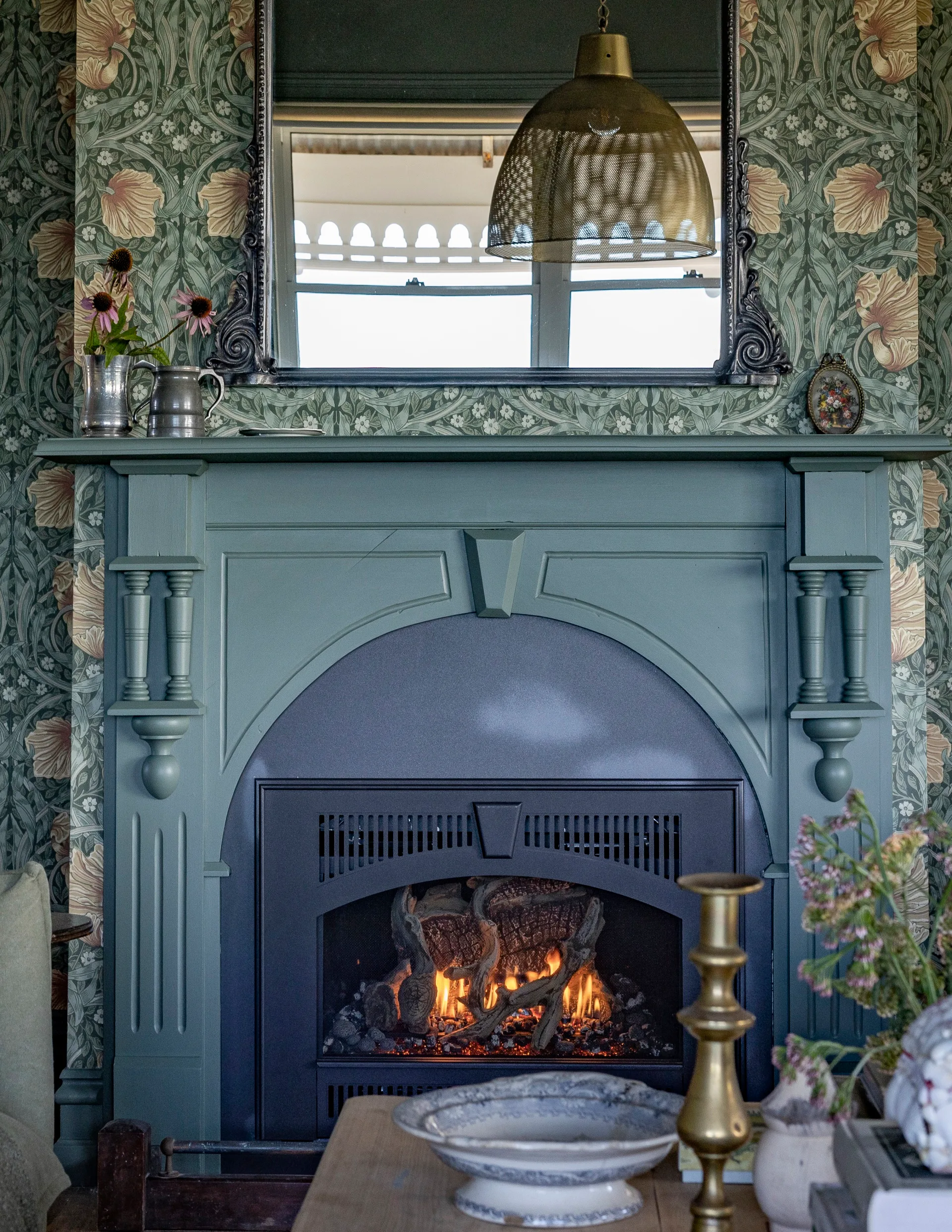 A fireplace with a painted green mantelpiece surrounded by dramatic patterned wallpaper