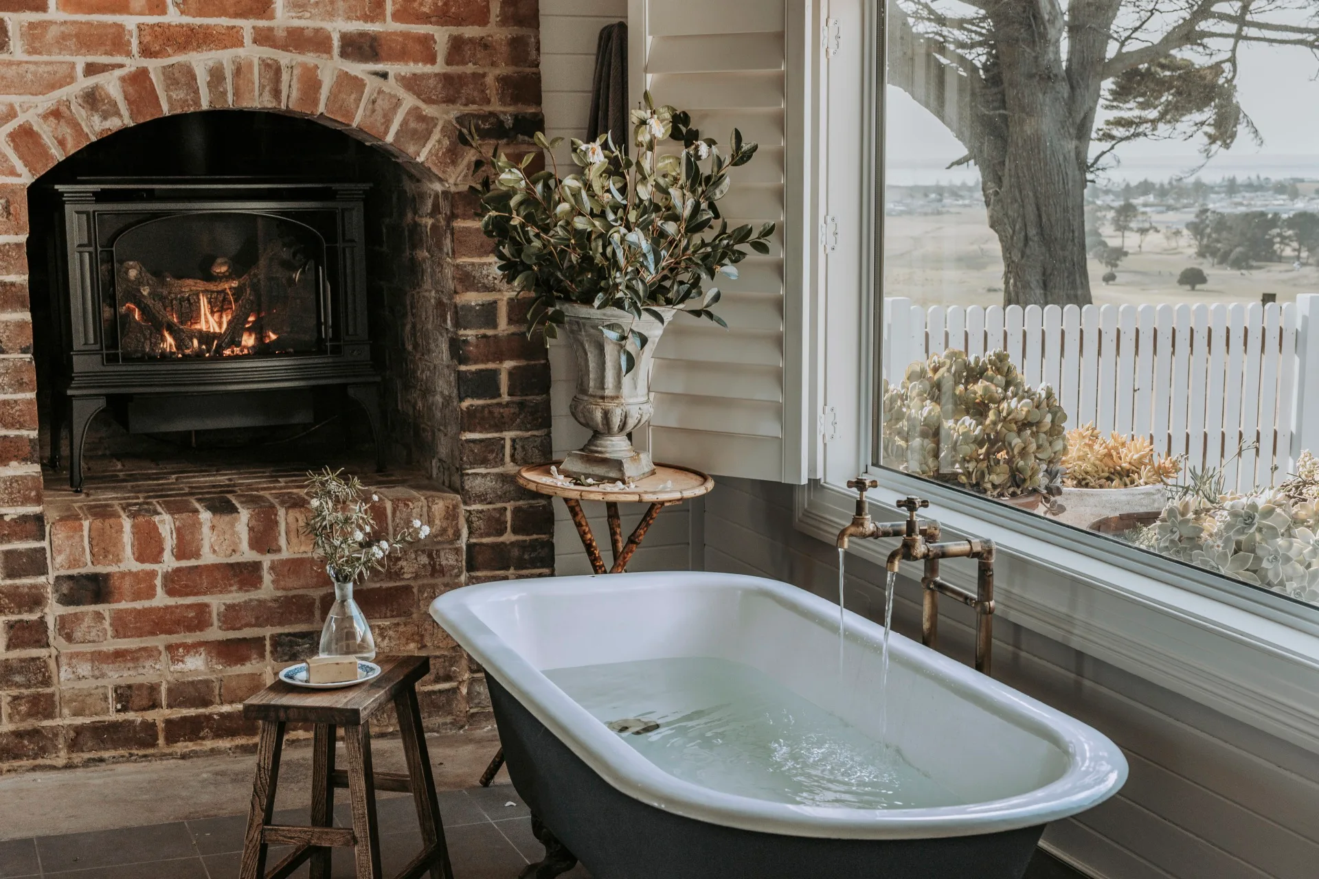 A freestanding, claw-foot bathtub sits alongside a large window with views out to the ocean, and an old fireplace.