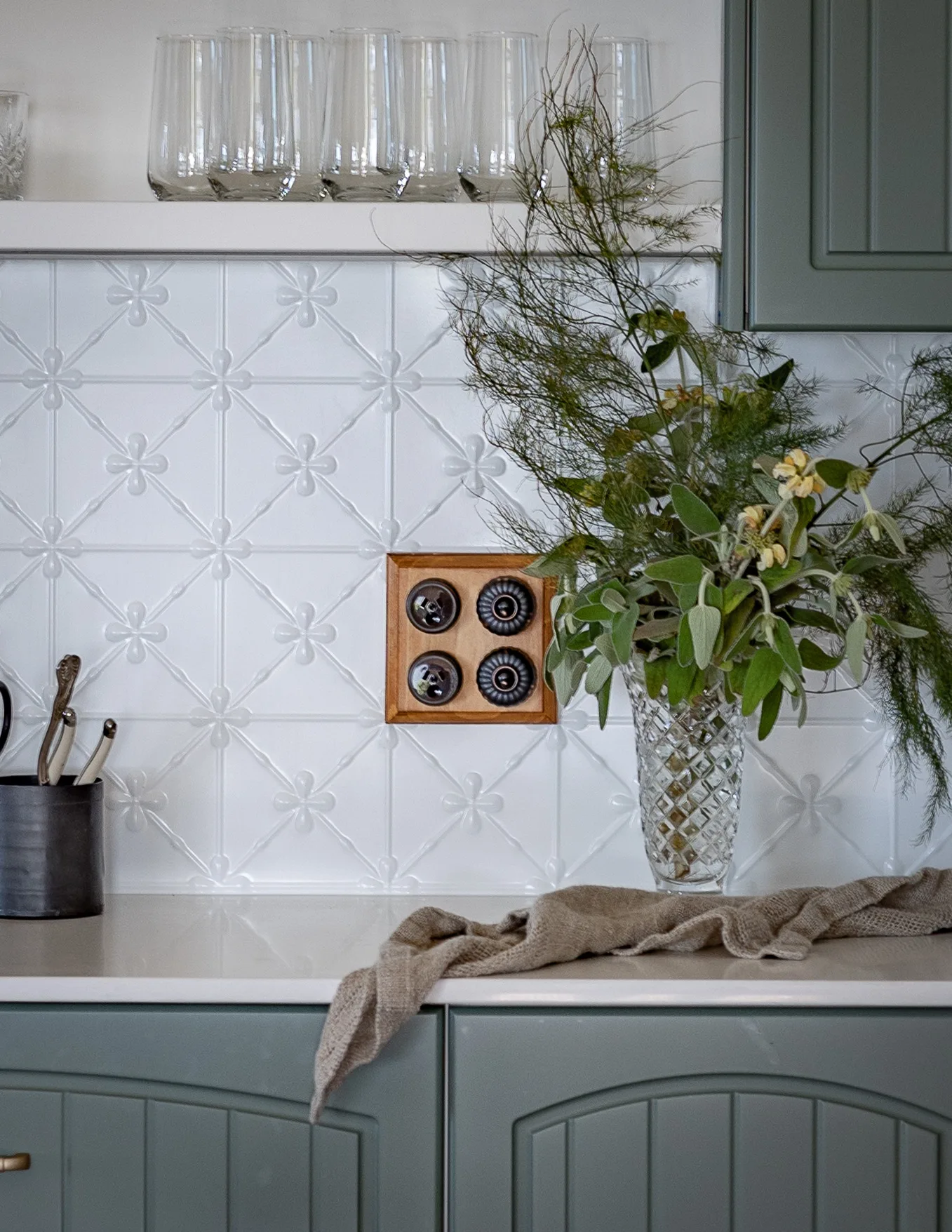 A kitchen with dark sage cupboards and a patterned, white metal splashback