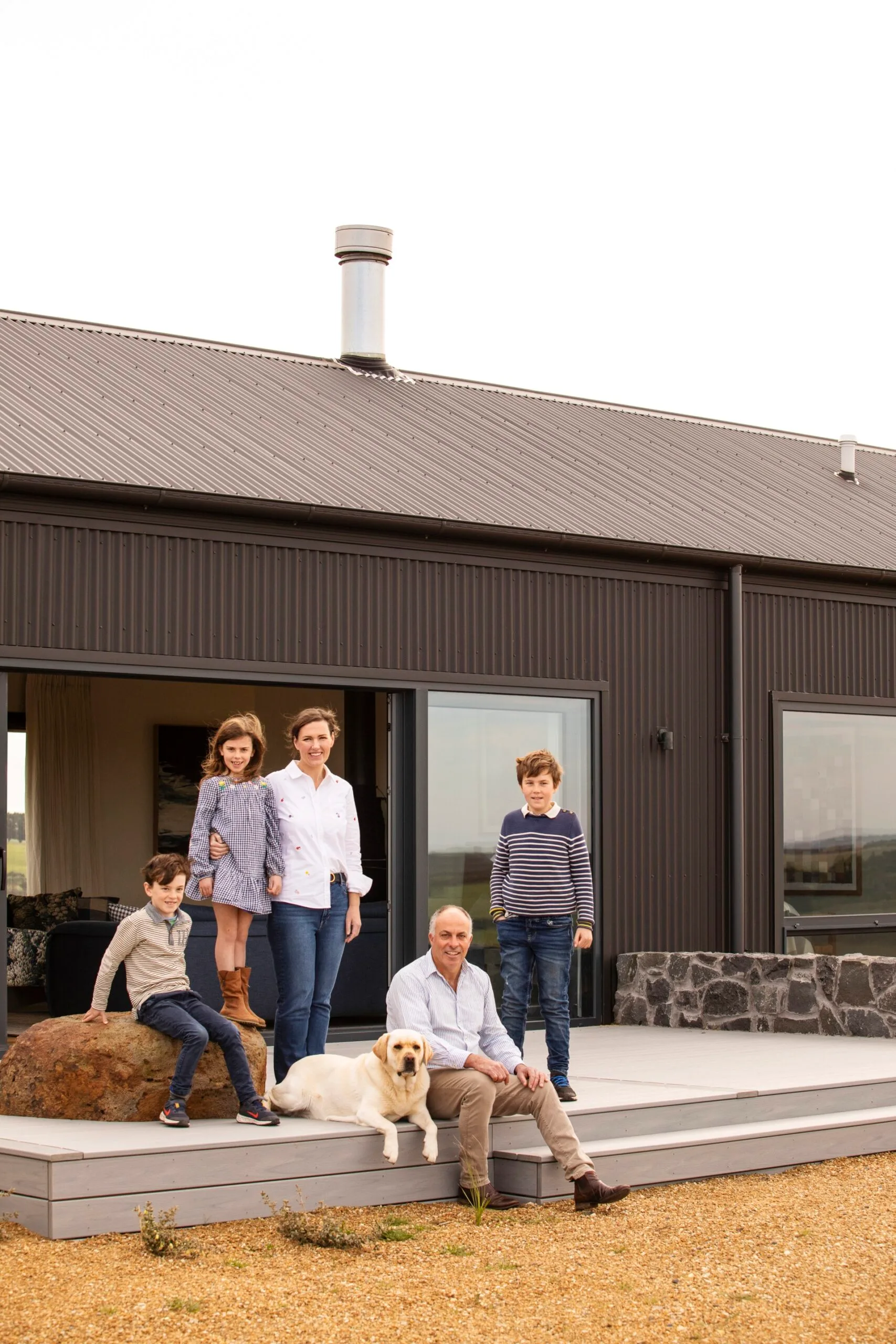 A young family stand outside a black house with a corrugated exterior.