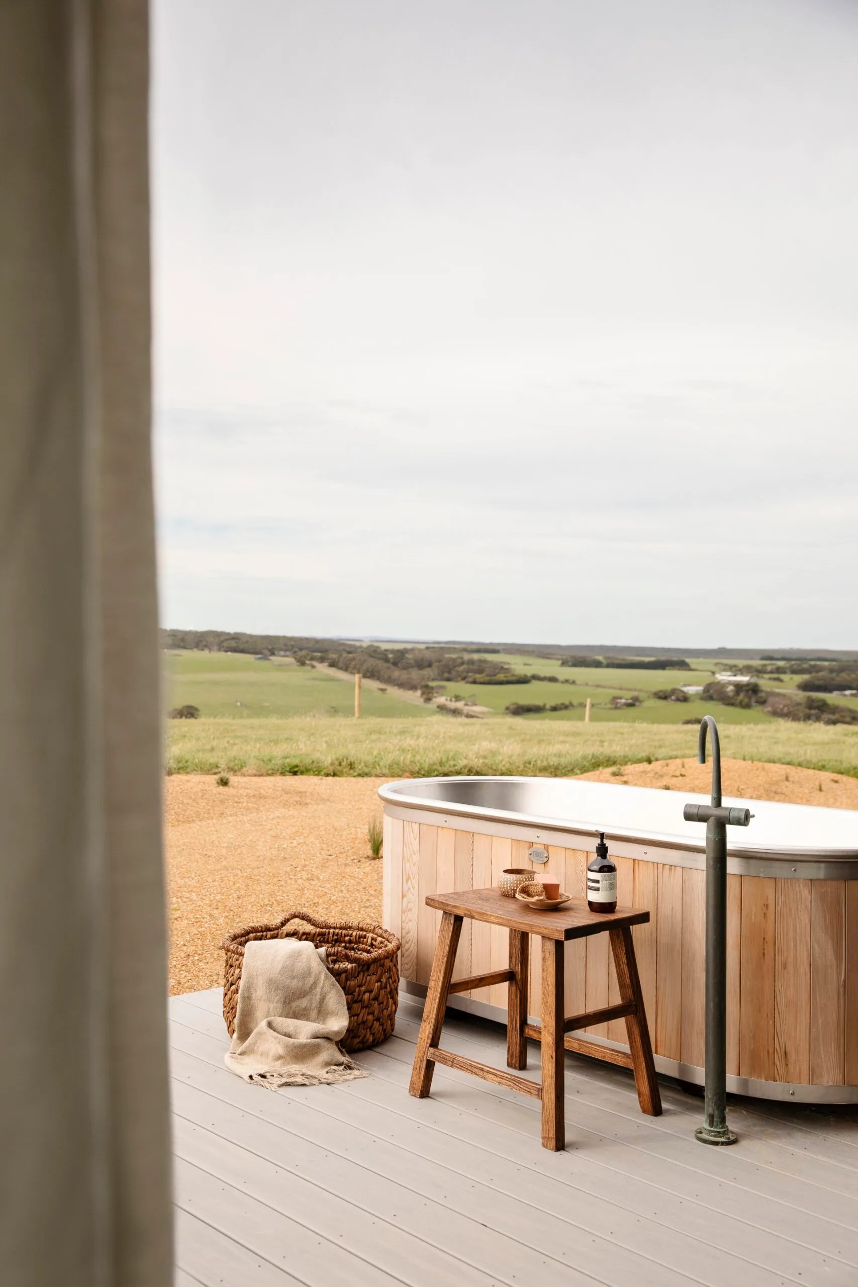 An outdoor wooden bath on a deck with a view of rolling hills and paddocks in Victoria, Australia.