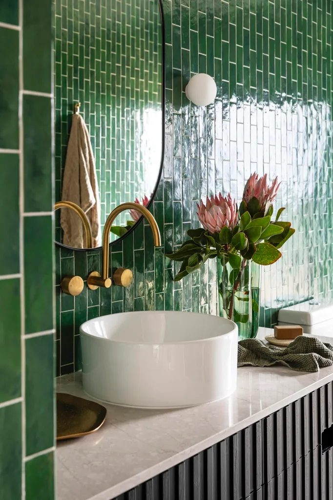 A bathroom vanity with shiny, bright green tiles, an oval-shaped mirror and gold hardware.