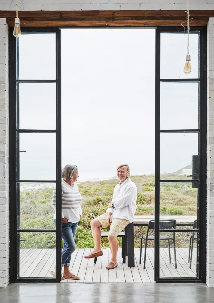 Double-height black metal doors look out to a coastal view where a couple stand on the balcony