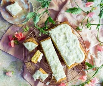 A lime and coconut slice with white icing sits on a wire rack to cool, surrounded by flowers