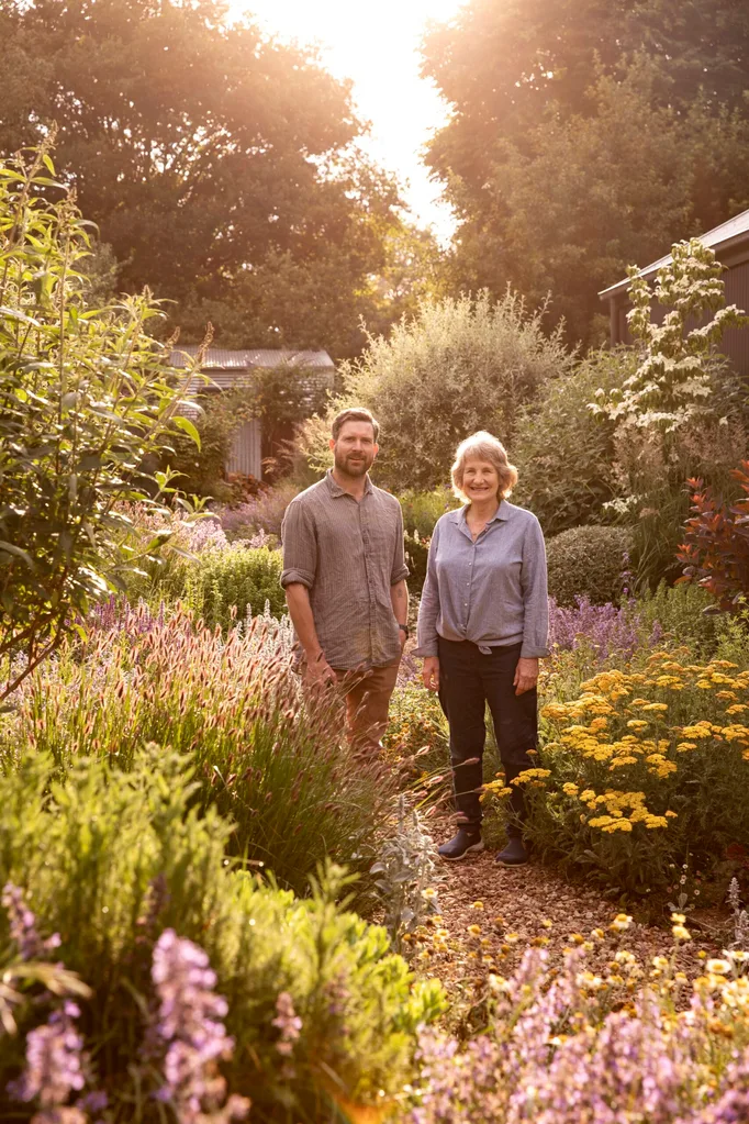 A man and a woman stand in a wild garden surrounded by flowers and plants