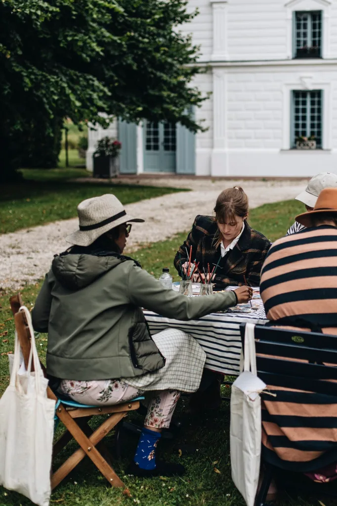 Women sit at a table painting with a French-style chateau in the background.