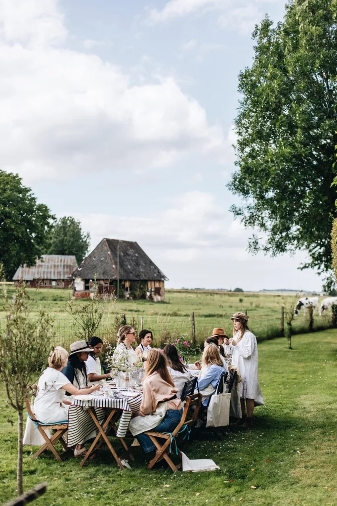 A group of women sit at a table in a garden in Normandy. In the background is a charming rustic cottage.