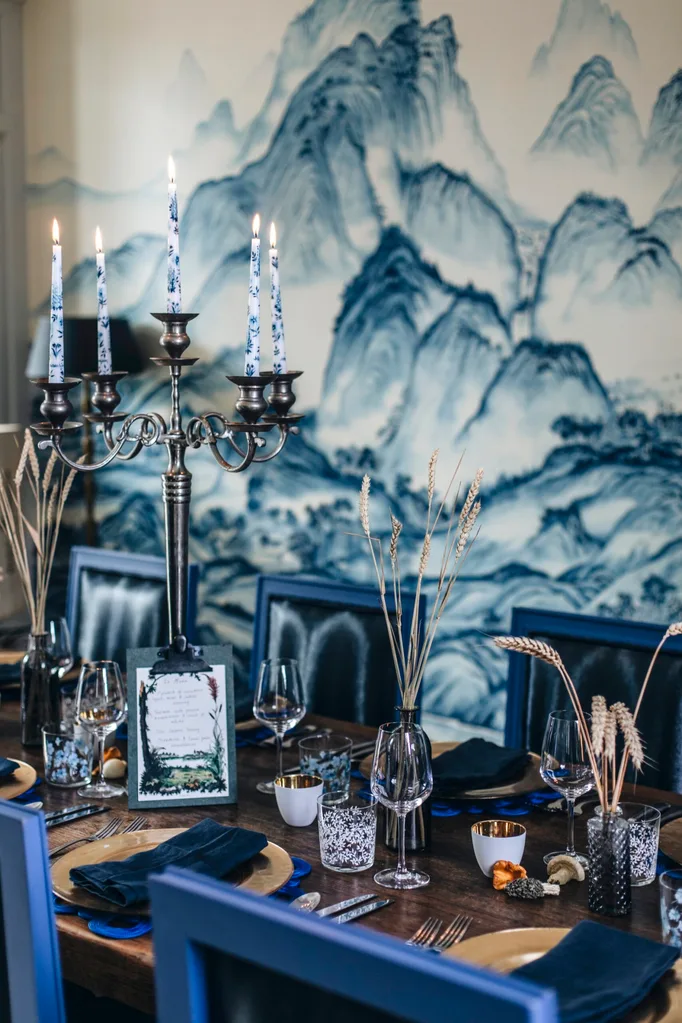 A beautiful wooden table with blue chairs, candles and napkins. The wall in the background is covered in blue and white wallpaper.