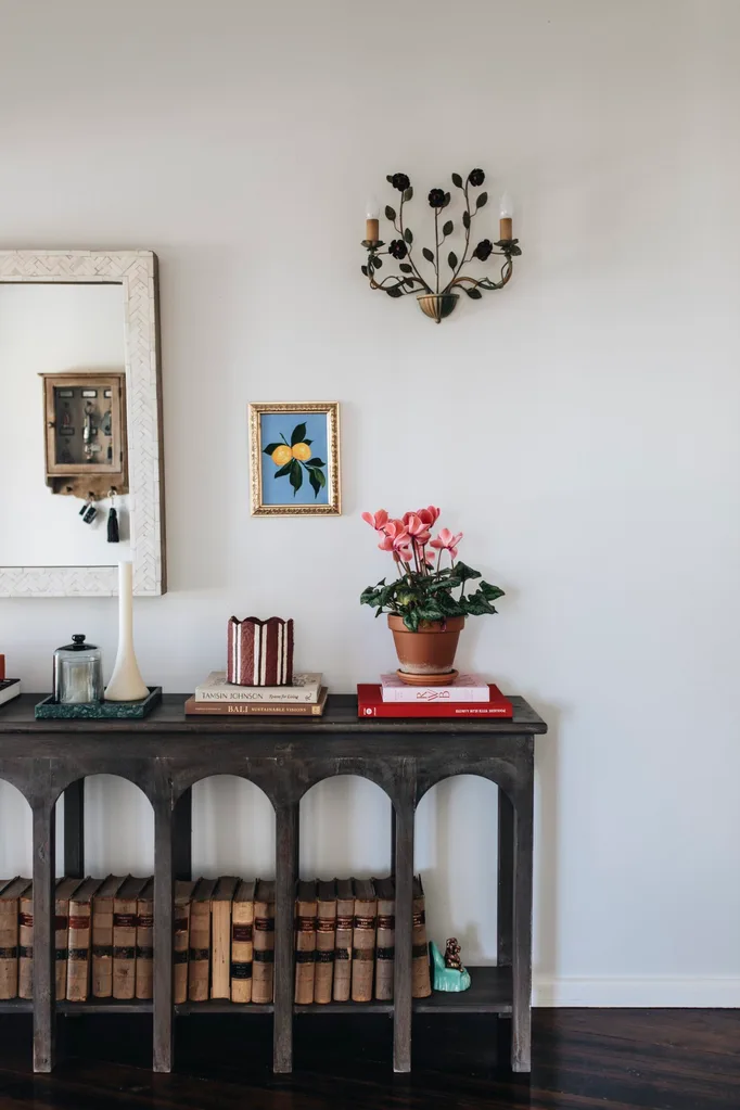 A console table with books and plants sits against a white wall decorated with a large mirror, a small lemon painting and a light sconce