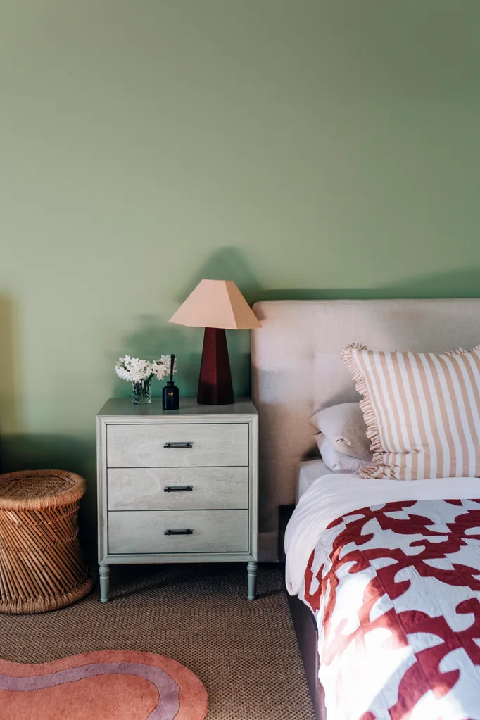 A bedroom with a dusty green wall, pink and red bedding and a sisal rug on the floor