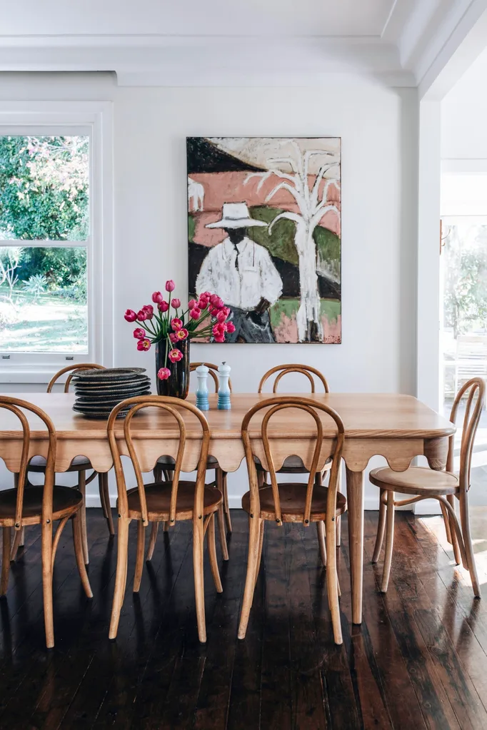 A light-filled dining room with an oak dining table and chairs. A large painting hangs on the white wall in the background and a vase of pink tulips sits on the table.