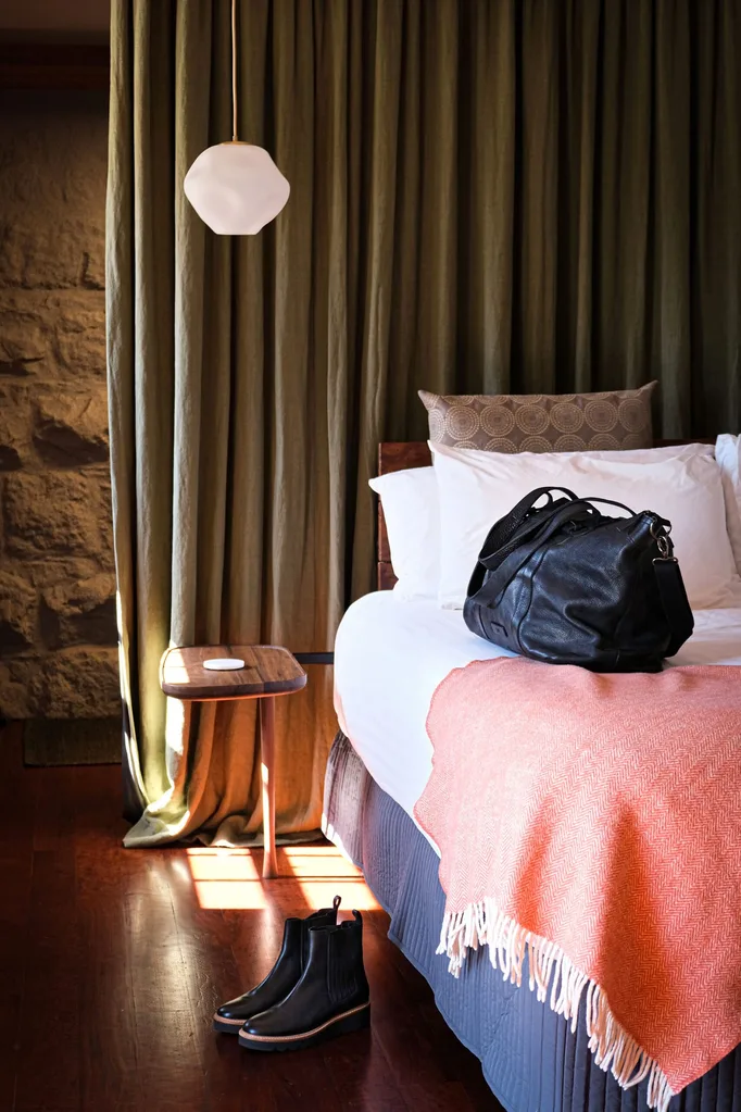 Bed with white bedlinen and pink throw with a heavy green curtain behind it, and a stone wall behind that in a travel story about a Grampians hotel