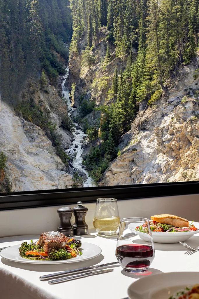 View through train window of steep rocky gorge with stream; inside the train, the table is set for dinner with wine and plates of food in Travel Canada Rocky Mountains
