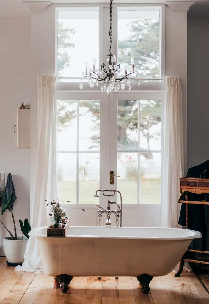 Freestanding white clawfoot bath in front of French doors out to garden, with chandelier overhead in a Grand Ridge House tour