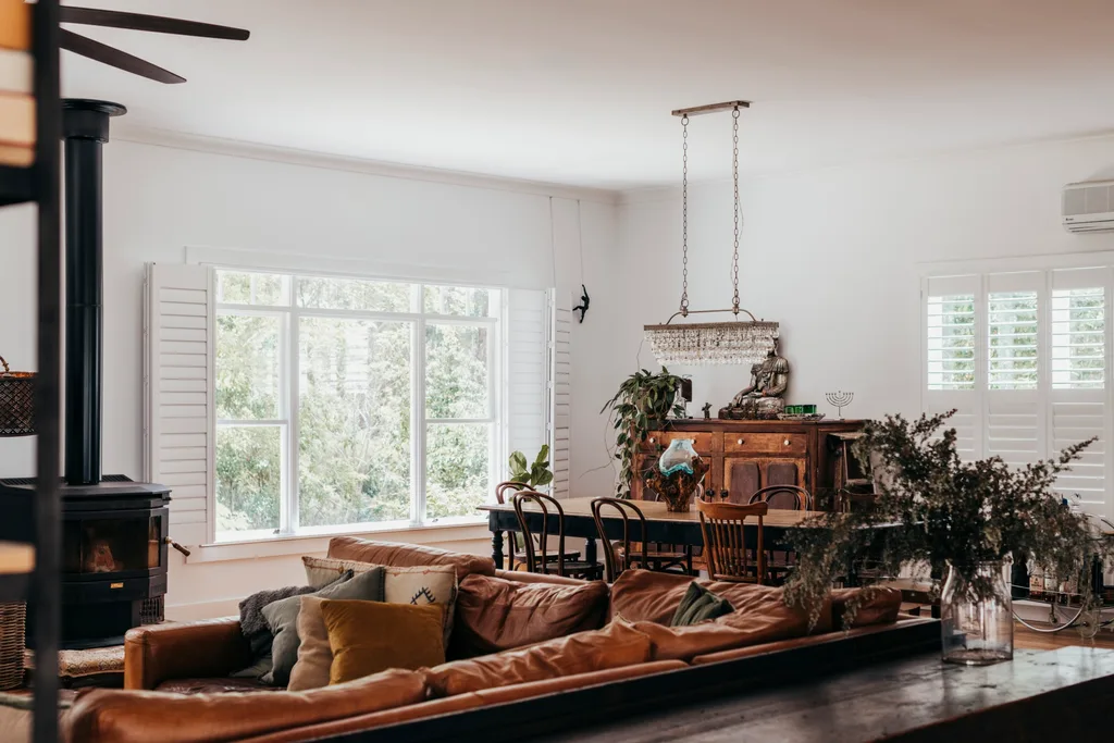 White-painted living-dining room with brown leather corner sofa, timber dining table and mismatched dining chairs with low-slung rectangular chandelier above, and timber sideboard with plants and a Buddha statue on it