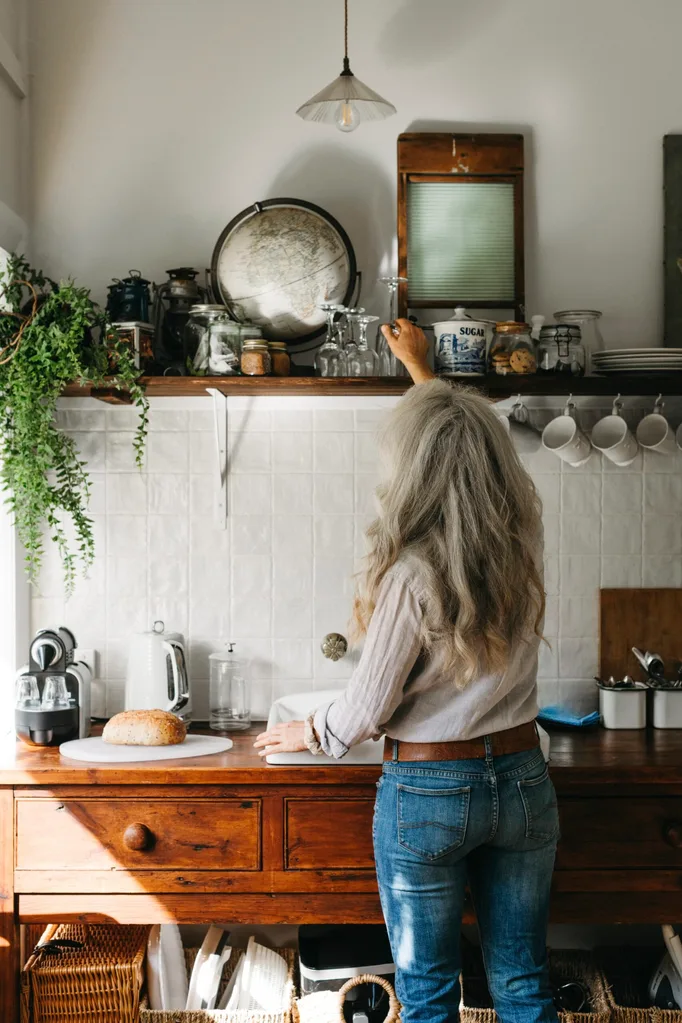 Woman in kitchen with timber lower cupboards, white-tiled splashback and open shelf above with mugs, glasses, plants and a globe on it