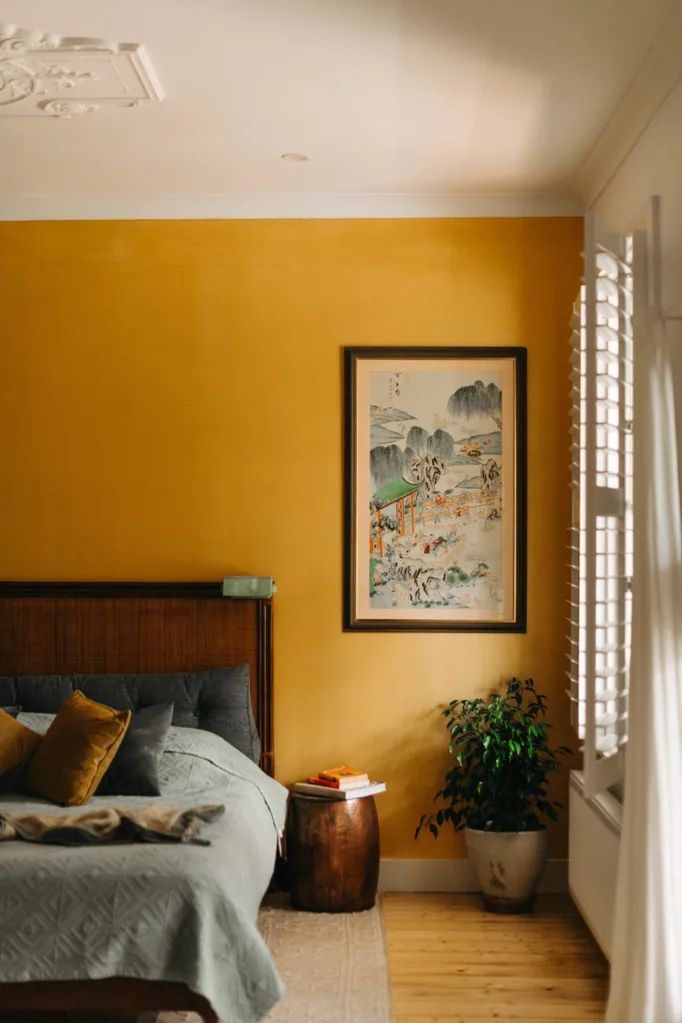 Bedroom with mustard-painted walls, rattan bedhead and grey bedlinen, brass stool for a bedside table, large Japanese print on wall, potted plant in corner, white platnation shutters