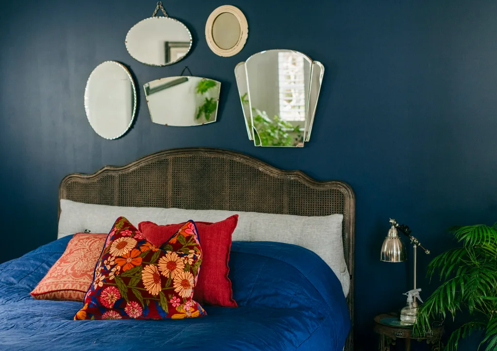 Blue-painted bedroom with vintage mirrors above the bed, blue bedlinen and mismatched red and yellow cushions on the bed in a Grand Ridge House tour
