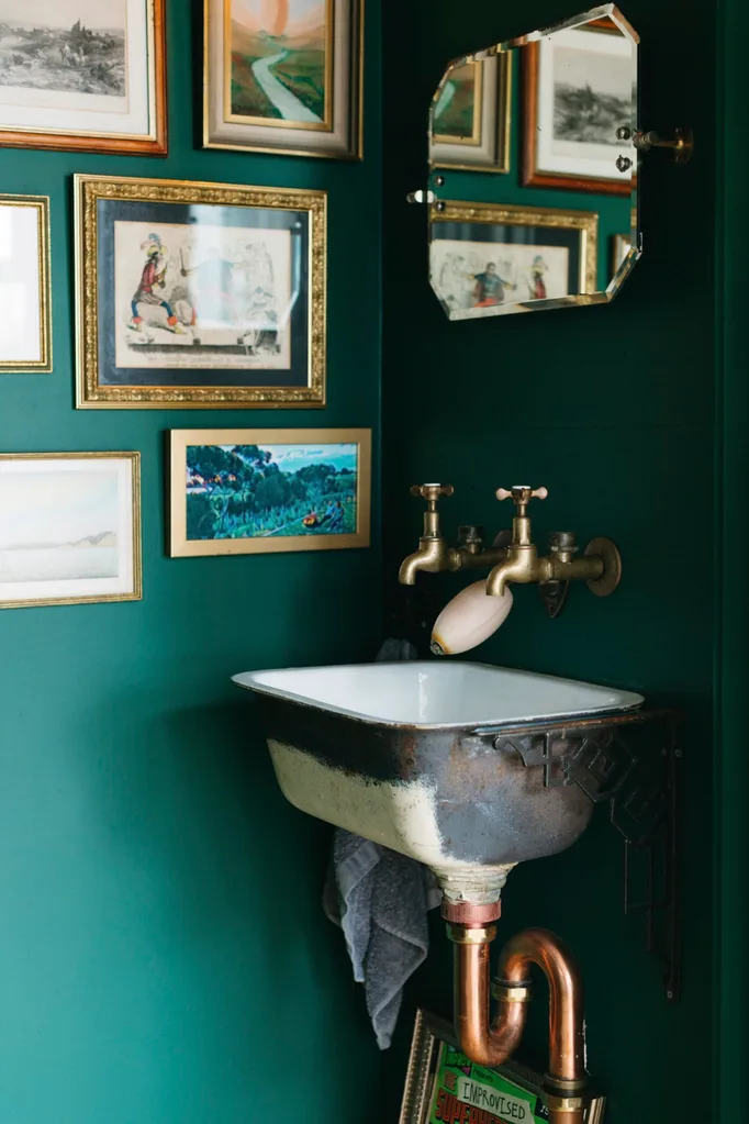 Forest green-painted bathroom with salvaged metal sink and brass taps, and lots of gilt-framed pictures on walls