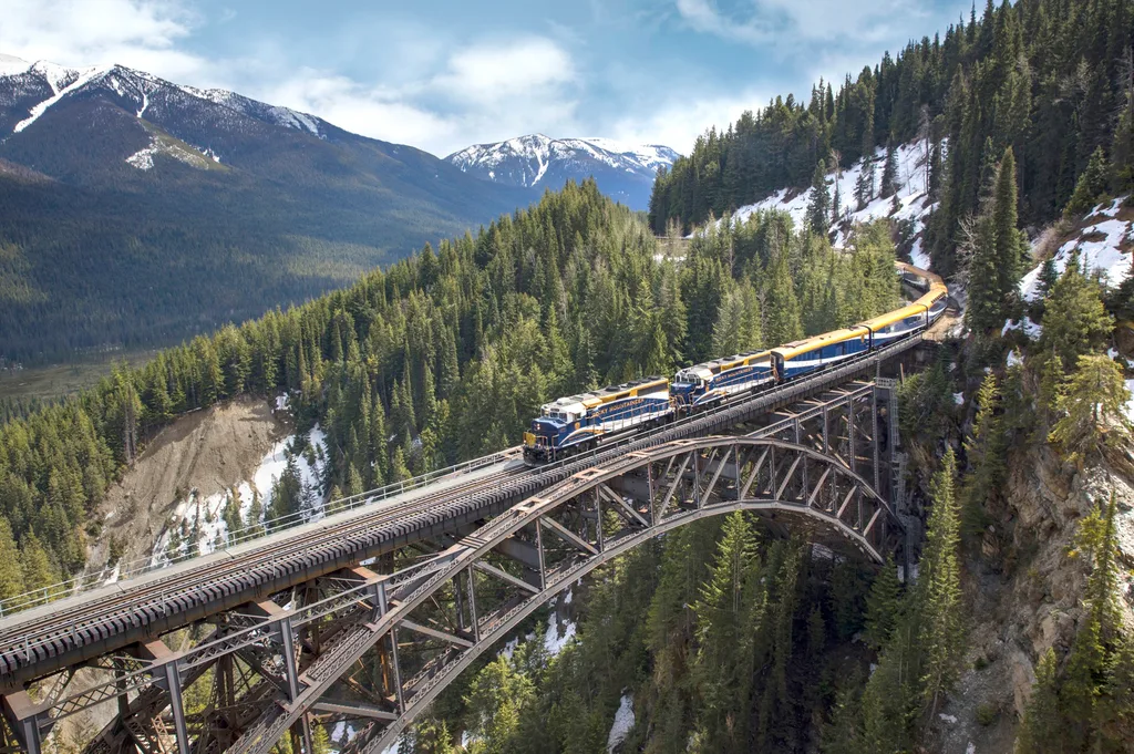 Train running on bridge over steep valley with fir trees and patches of snow; snow-capped mountains behind in Travel Canada Rocky Mountains