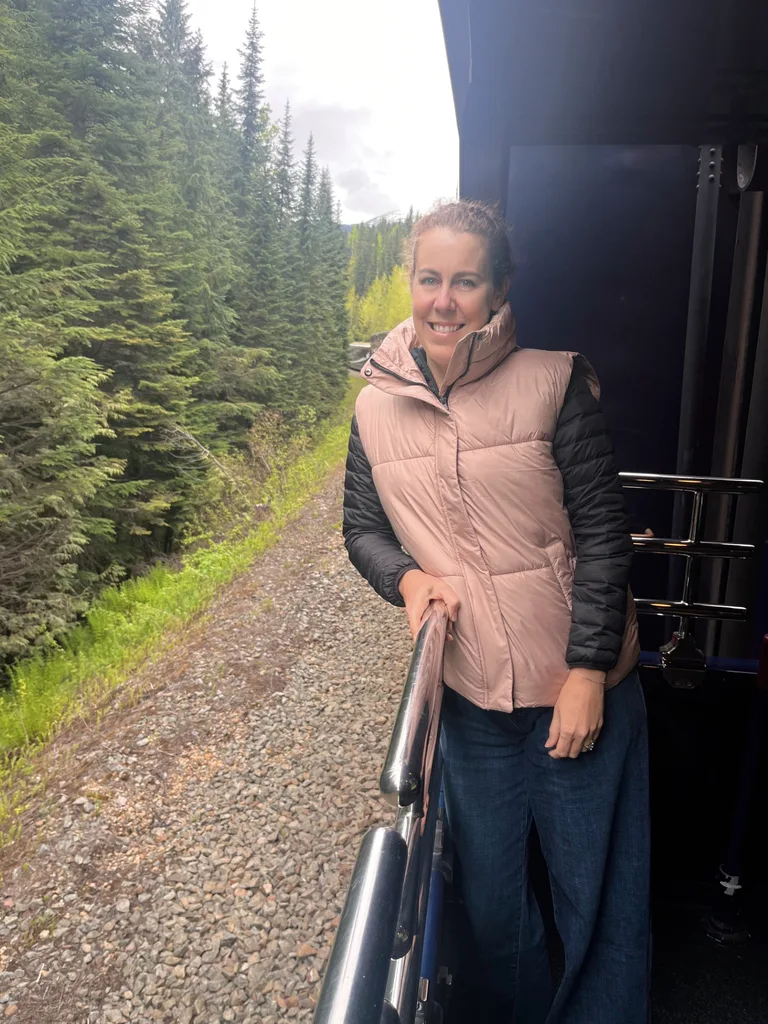 Woman in puffer jacket leans out of train going along valley lined with fir trees