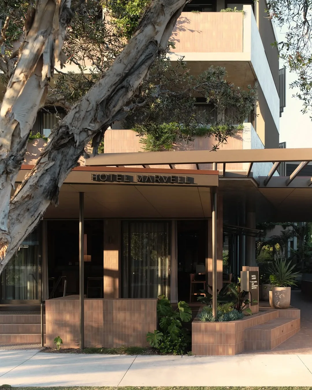 A brick building with three-levels, garden beds, a sign that says "hotel marvell" and a tree trunk