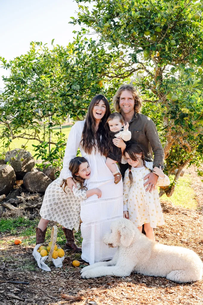 A family with three young daughters stands beside a lemon tree with a dog at their feet.