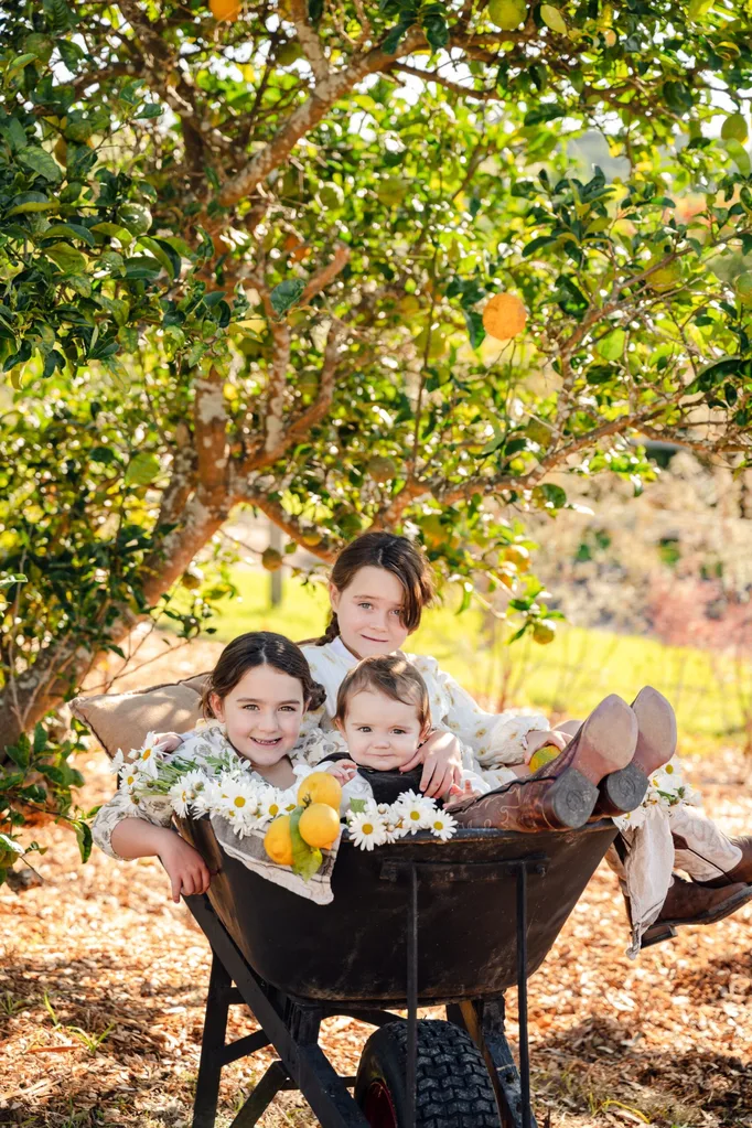 Three young girls sit inside a wheelbarrow under a citrus tree.