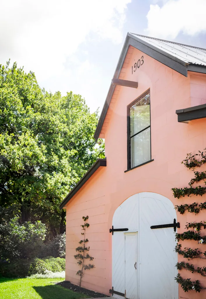 A pink building with black timbers and white arched double doors has the date 1903 painted at its peak and two trees espaliered on either side of the door. There's a row of trees behind.