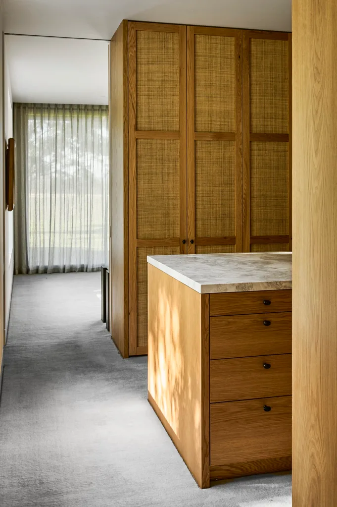 A walk-in wardrobe with a large, marble topped island and floor-to-ceiling wooden cupboards with cane webbing detailing.