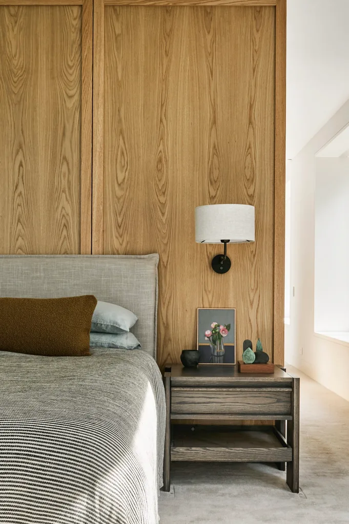 A bedroom with a custom joinery and wall panelling in oak veneer, a grey upholstered headboard, and a striped, black and white coverlet.