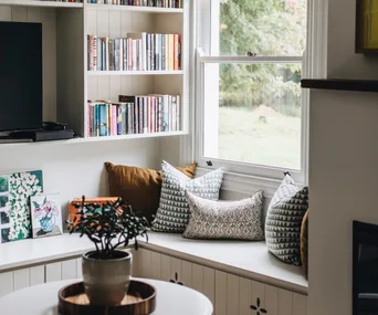 A window seat with cushions below a shelf filled with books