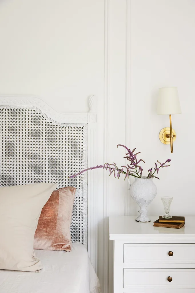 Bedroom with white walls, a rattan bedhead, a white bedside table and a traditional wall light.