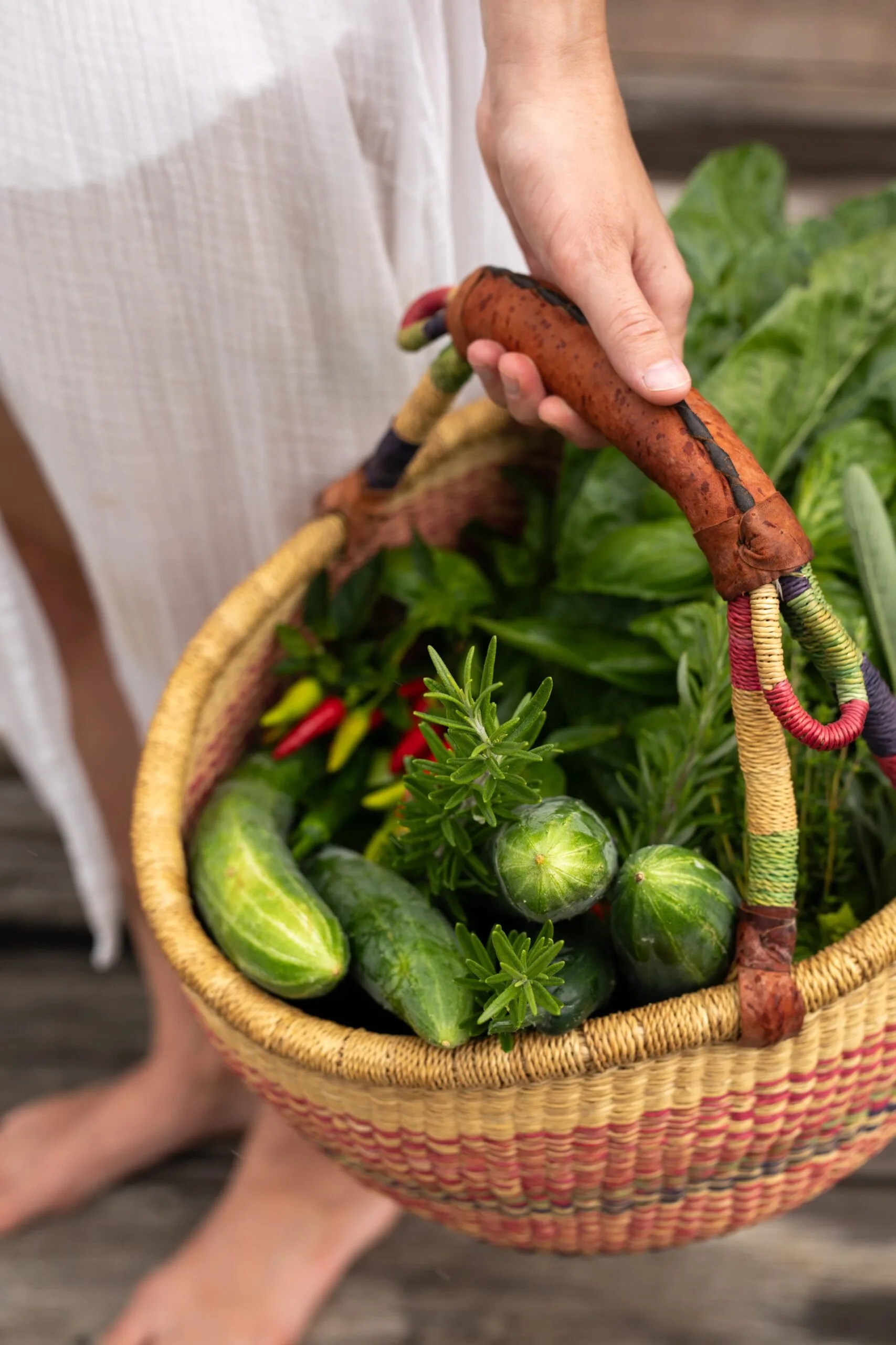 A person holding a woven basket full of fresh vegetables and herbs