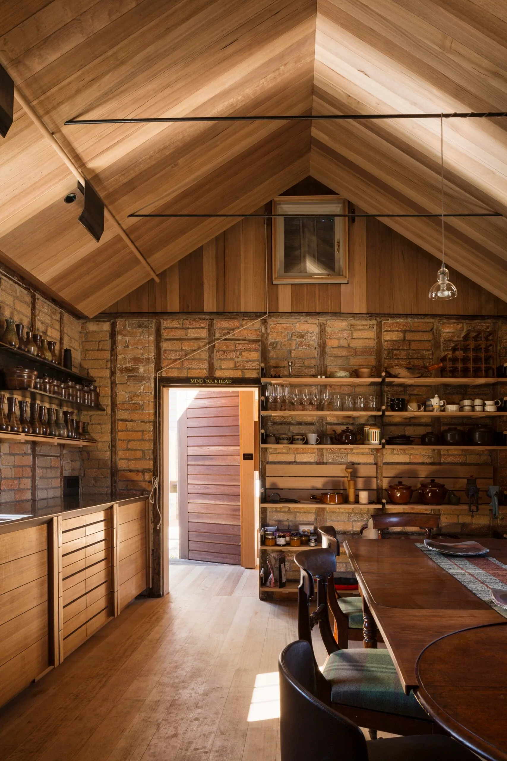 A timber kitchen and dining area with a pitched roof and shelving lined with crockery and glassware