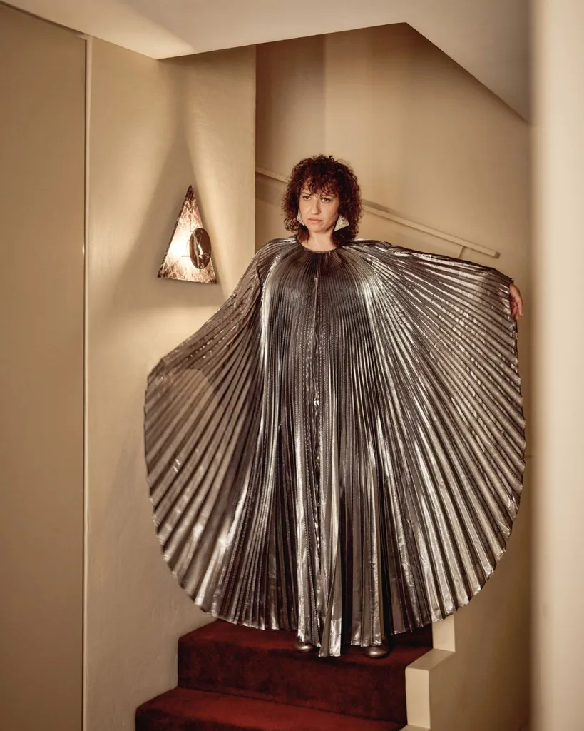 Anna Viniero stands in the staircase of her Sydney home. She wears a pleated metallic cape dress.