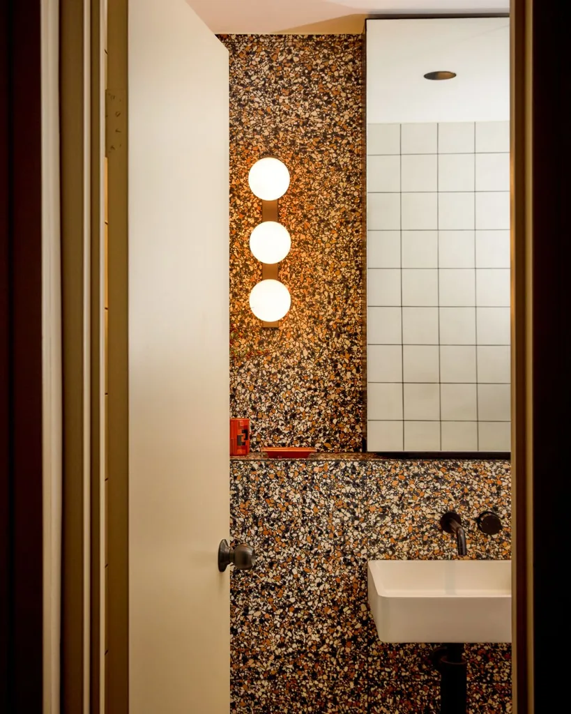 A doorway opens into a bathroom with floor to ceiling terrazzo tiles, a vanity mirror and single sink with brass tapware.