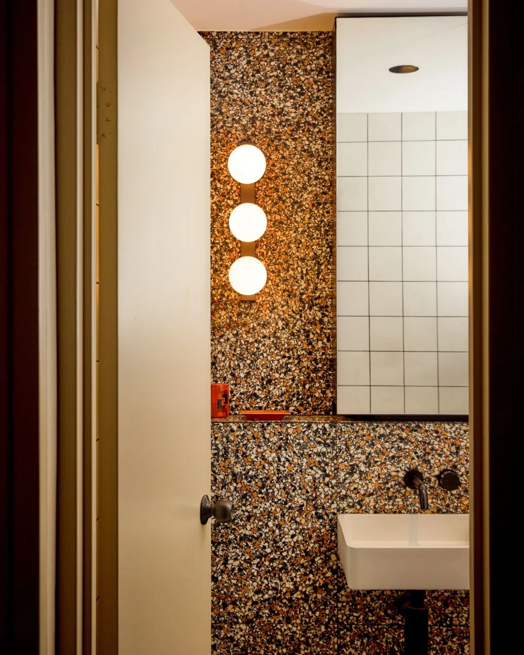 A doorway opens into a bathroom with floor to ceiling terrazzo tiles, a vanity mirror and single sink with brass tapware.